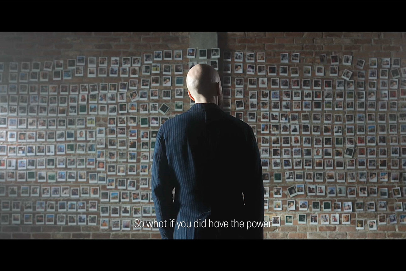 Derren Brown stands with his back to the camera stands and stares at a lrage wall of polaroid pictures.