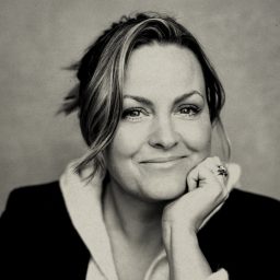 Jo Joyner smiling, wearing a black top, with a blurred grey background.