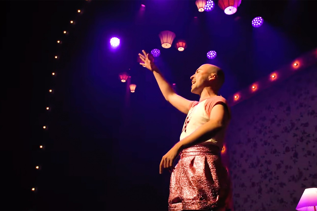Rob Madge performs in pink skirt and white top gestures on stage under purple lighting and hanging lanterns.
