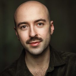 Rob Madge, wearing a dark shirt, smiles softly against a dark, blurred background.