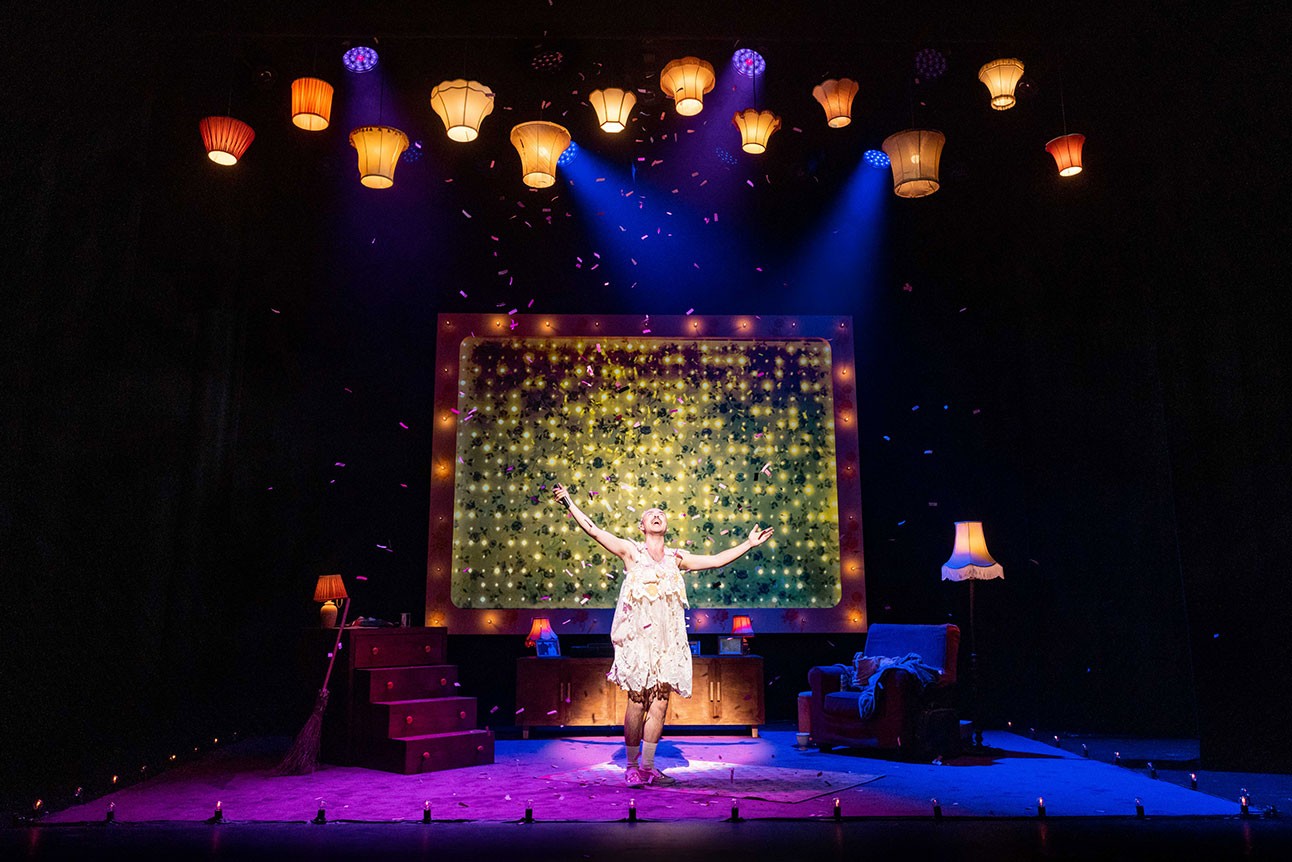 Performer on stage with arms raised, surrounded by colourful lights and vintage lampshades.