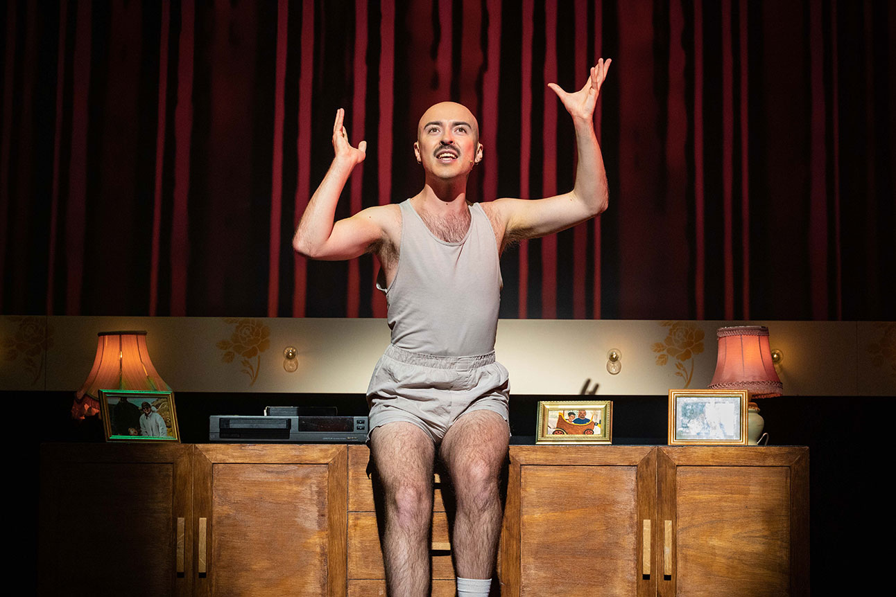 Rob Madge performs energetically on stage, wearing a grey outfit, with a red curtain backdrop.