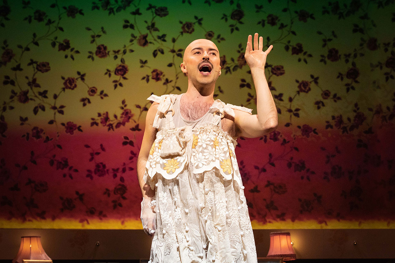 Rob Madge performs energetically on stage, wearing a white lace dress, floral backdrop behind.