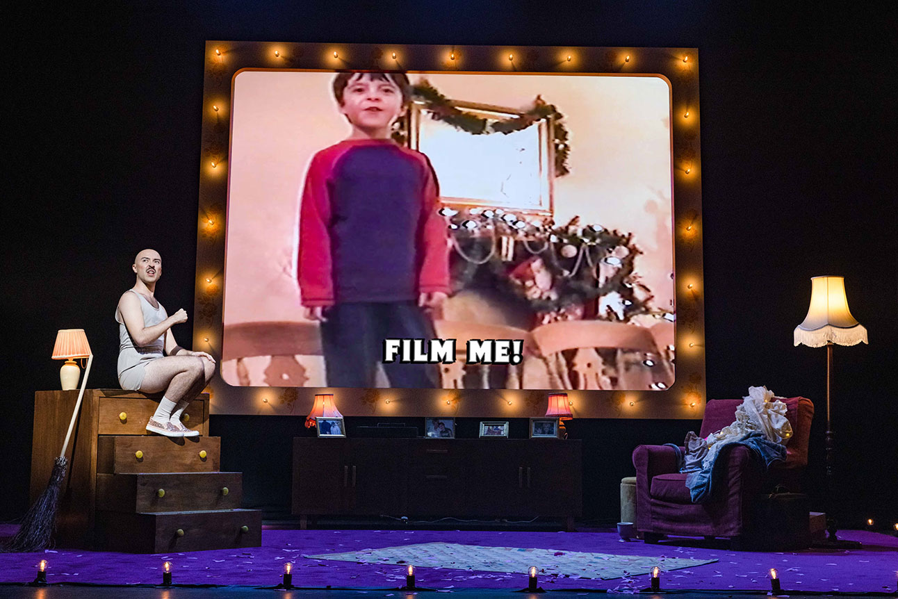 Stage with large screen showing child; text reads "FILM ME!". Two seated figures on stage.