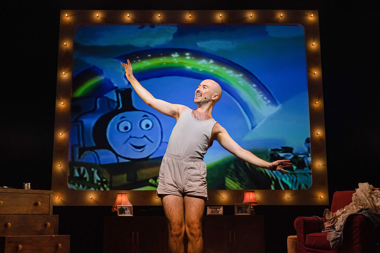 Rob Madge performs on stage in front of a Thomas the Tank Engine backdrop, wearing grey attire.