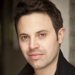A man with short dark hair and a neutral expression, wearing a dark jacket, indoors.