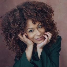 Smiling woman with curly hair rests chin on hands, wearing a dark green top, brown background.