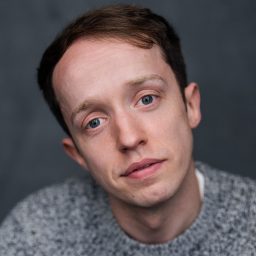 A person with short brown hair, wearing a grey jumper, against a dark grey background.