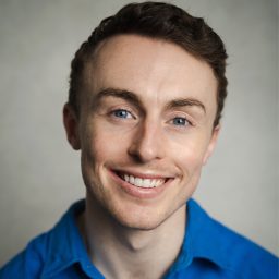 Smiling man in blue shirt against neutral background, short brown hair, light complexion.