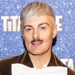 A man with short grey hair and moustache, wearing a brown jacket and white bow tie.