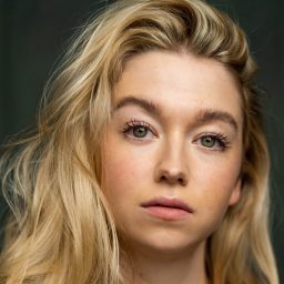 A woman with long blonde hair and a neutral expression against a dark background.