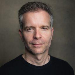 Damian Humbley headshot; neutral expression, short grey hair, wearing a black tshirt, grey background.