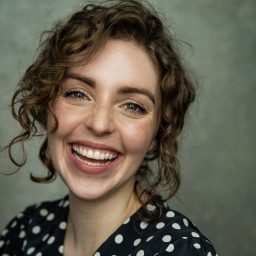 Smiling woman with curly hair, wearing a polka dot top, against a grey background.