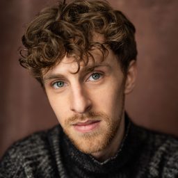 A man with curly brown hair and a beard, wearing a dark sweater, against a brown background.