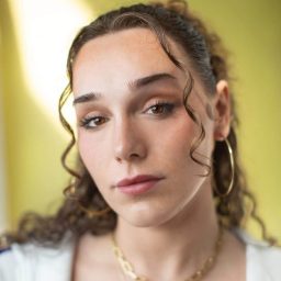 A person with curly hair and hoop earrings looks directly at the camera against a yellow background.