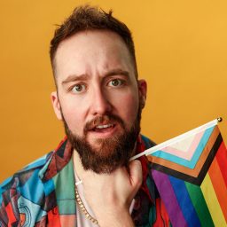 Jordan Paul Clarke holds a Pride flag against a yellow background, wearing a colourful shirt.