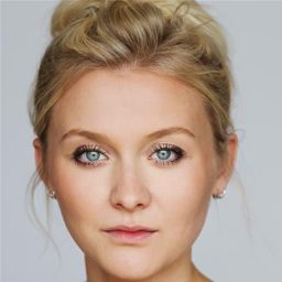 A woman with blonde hair in a bun, wearing earrings, against a plain grey background.