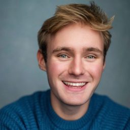 Smiling man with short blond hair wearing a blue jumper against a grey background.