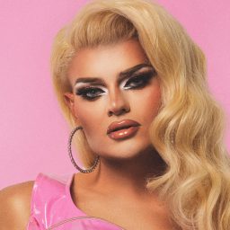 Person with long blonde hair, dramatic makeup, and hoop earrings against a pink background.