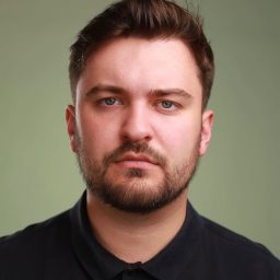 A man with short brown hair and a beard, wearing a dark shirt, neutral expression.