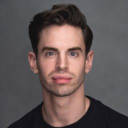 A man with short dark hair and a neutral expression, wearing a black shirt, grey background.