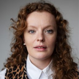 A woman with curly hair and a leopard print scarf, wearing a white shirt, neutral background.