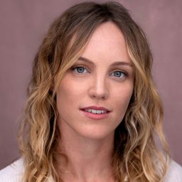 A woman with wavy blonde hair and a neutral expression against a soft pink background.