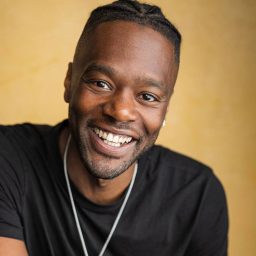 Smiling man with braided hair wearing a black shirt and necklace, against a warm yellow background.