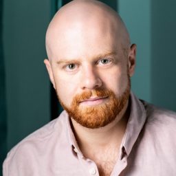A man with a bald head and red beard, wearing a light pink shirt, looks forward.