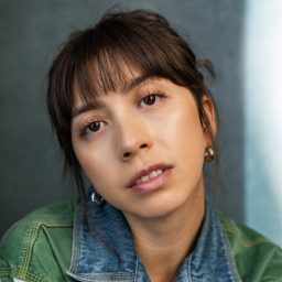 A woman with brown hair and fringe, wearing a green and denim jacket, gazes forward.