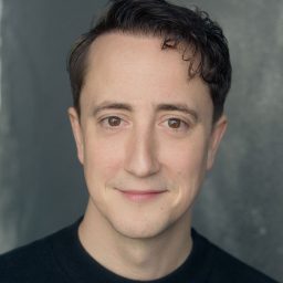 A man with short dark hair and a neutral expression against a grey background.