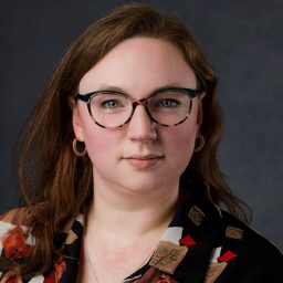 Micha Mirto with long brown hair, wearing glasses and a patterned shirt, against a dark grey background.