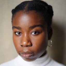 A person with braided hair and hoop earrings, wearing a white turtleneck, neutral background.