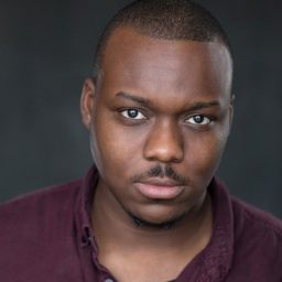 Raphael Bushay headshot; wearing a maroon shirt, neutral expression, dark background.