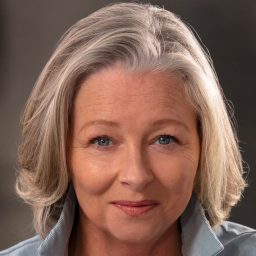 Sarah Ingram smiling, wearing a light blue shirt, with grey hair, neutral background.
