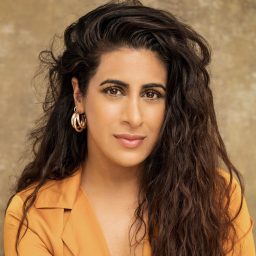 A woman with long, wavy hair wearing a mustard blouse and hoop earrings, neutral background.