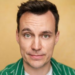 A man with short dark hair wearing a green striped shirt against a yellow background.