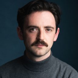 A man with dark hair and a moustache, wearing a grey turtleneck, against a dark background.