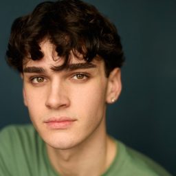 Young man with curly dark hair, wearing a green shirt, against a dark blue background.