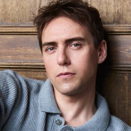 A man in a grey shirt leans against a wooden wall, looking directly at the camera.