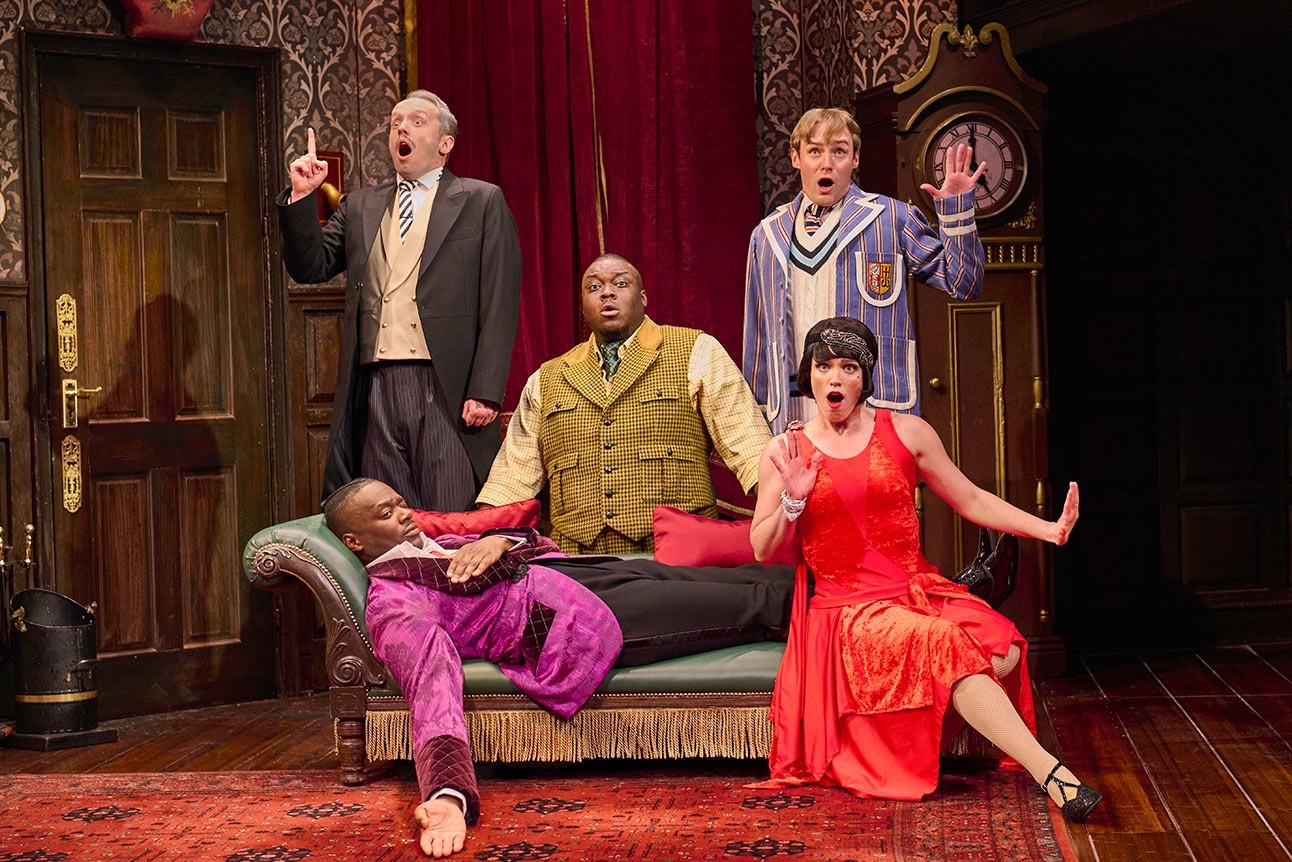 Five actors in theatrical costumes express surprise on a vintage set with red curtains and clock.