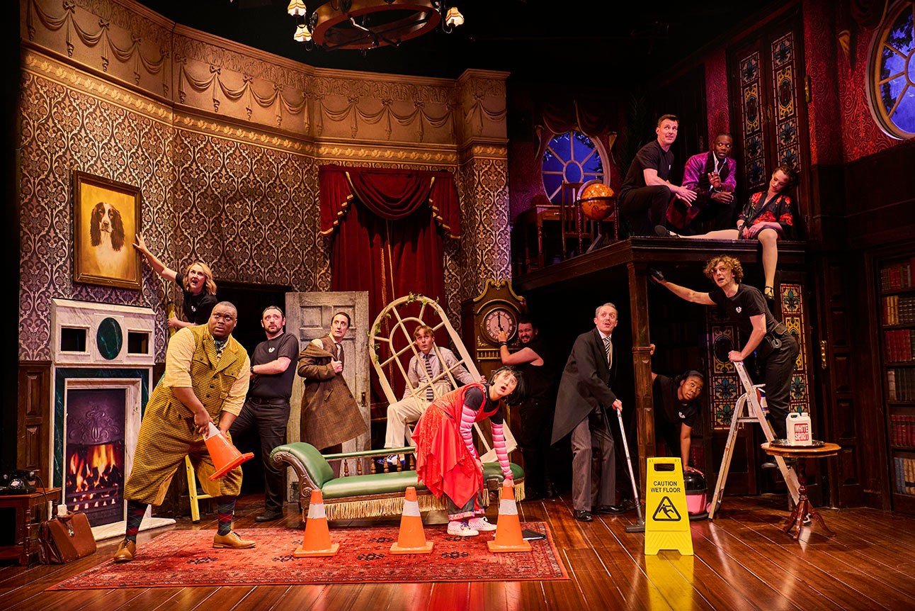 Theatre cast in comedic poses on stage with props, vibrant costumes, and ornate set design.
