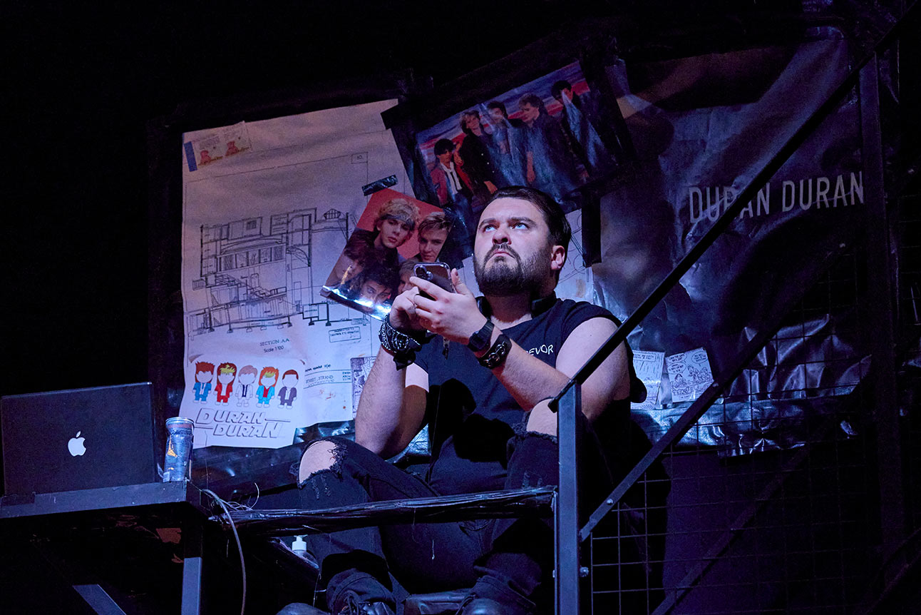 Man sitting on stage, holding a phone, surrounded by posters, wearing a black sleeveless shirt.