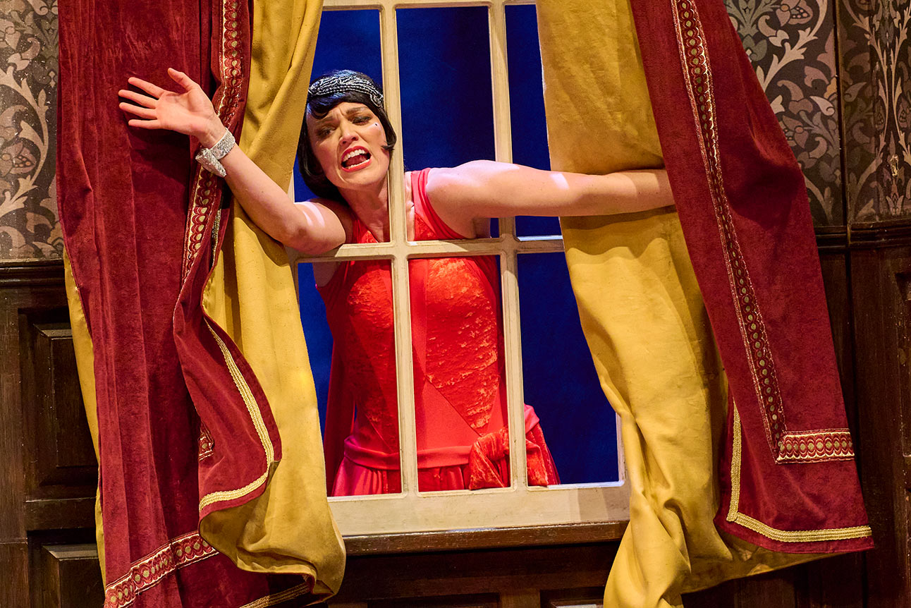 Woman in red dress leans out of window, framed by red and gold curtains, smiling.