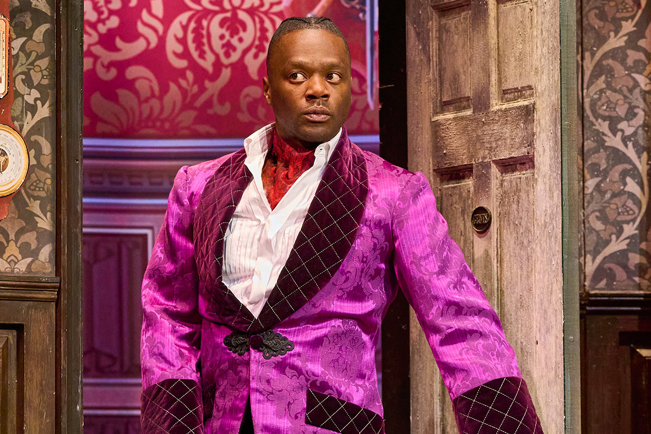 Person in vibrant purple patterned jacket stands in front of ornate pink wallpaper and wooden door.