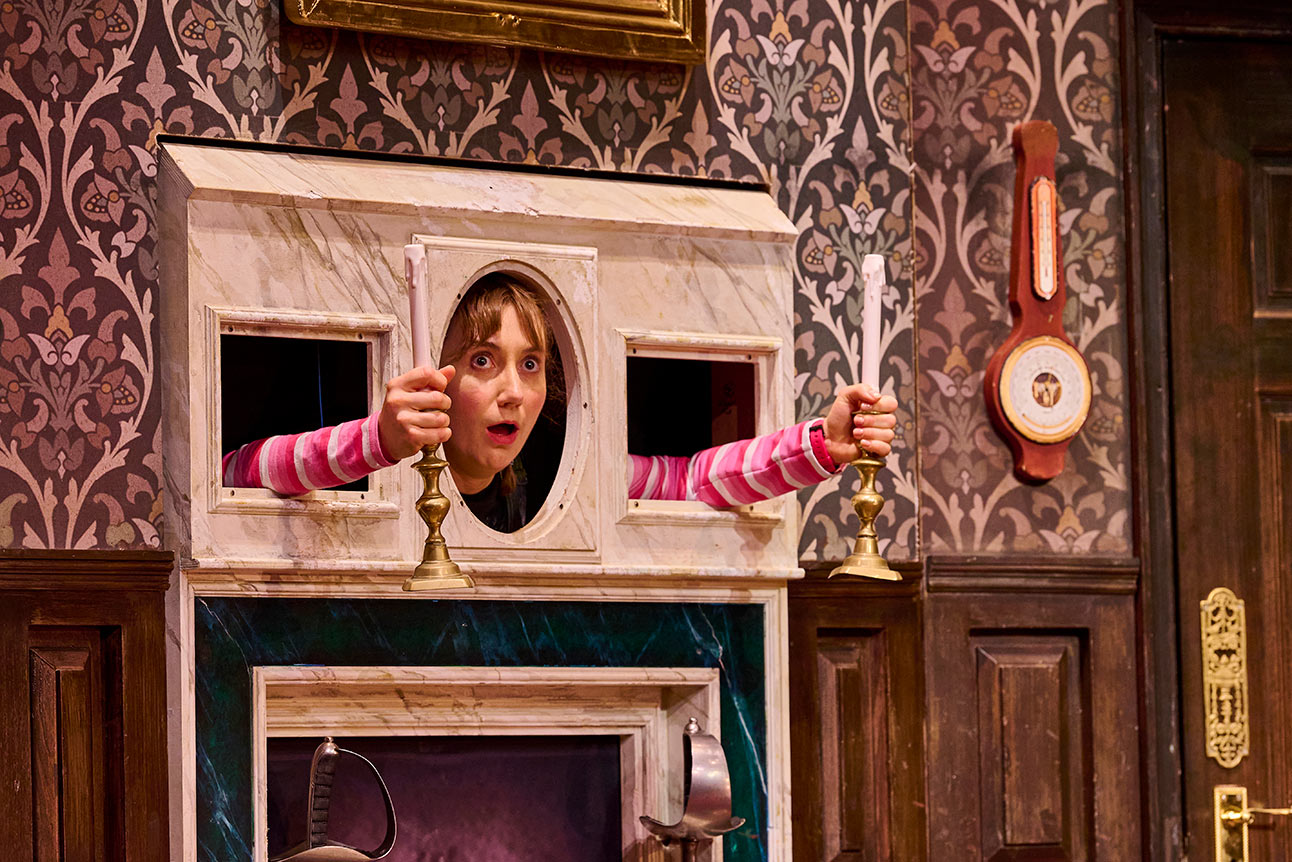 Person in striped top peers through fireplace, holding candlesticks; ornate wallpaper and clock in background.