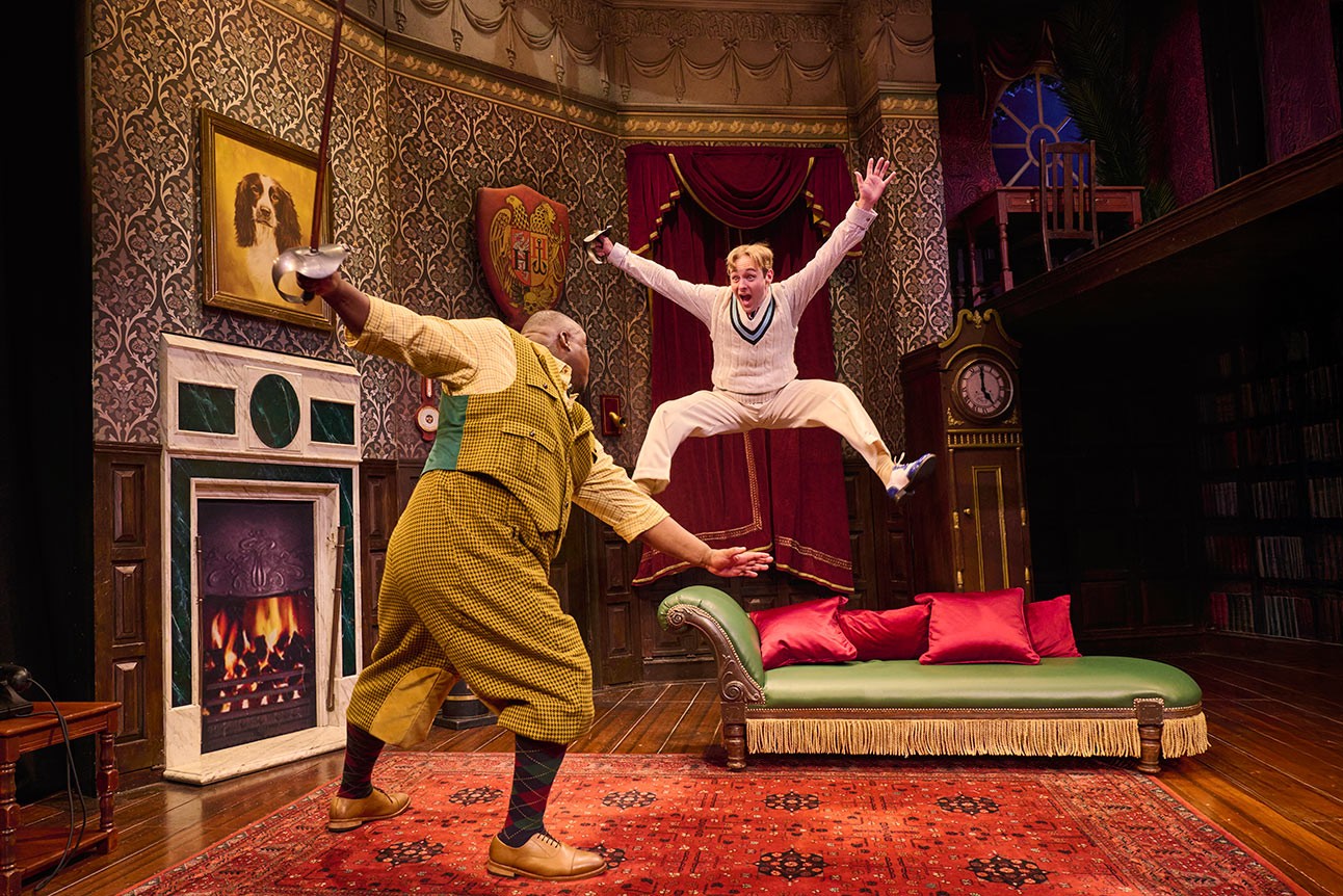 Two actors perform energetically on stage; one leaps in mid-air, the other gestures dramatically.