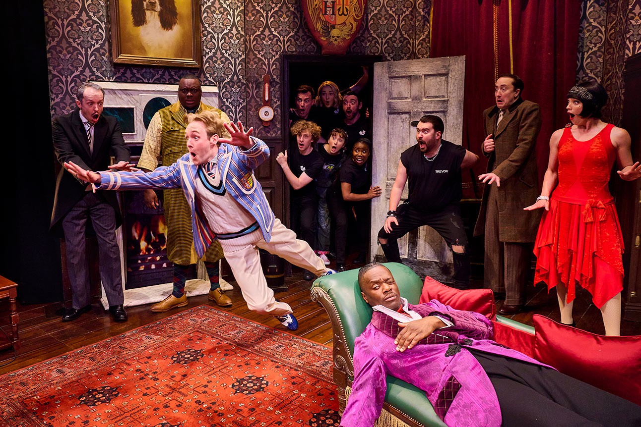 Actors in colourful costumes perform energetically on a detailed stage set with ornate wallpaper and props.