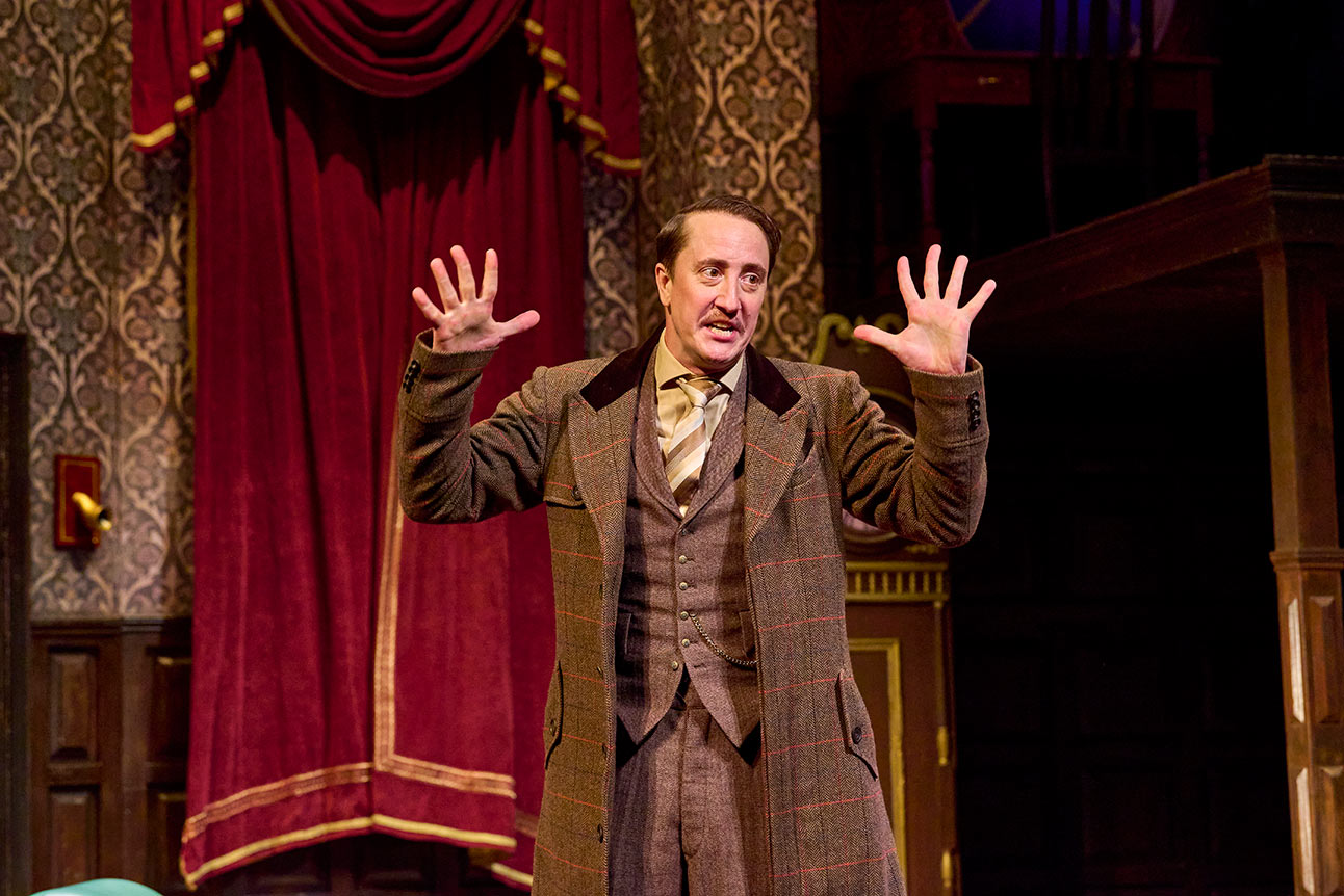 A man in a brown suit gestures with raised hands on a theatre stage with red curtains.
