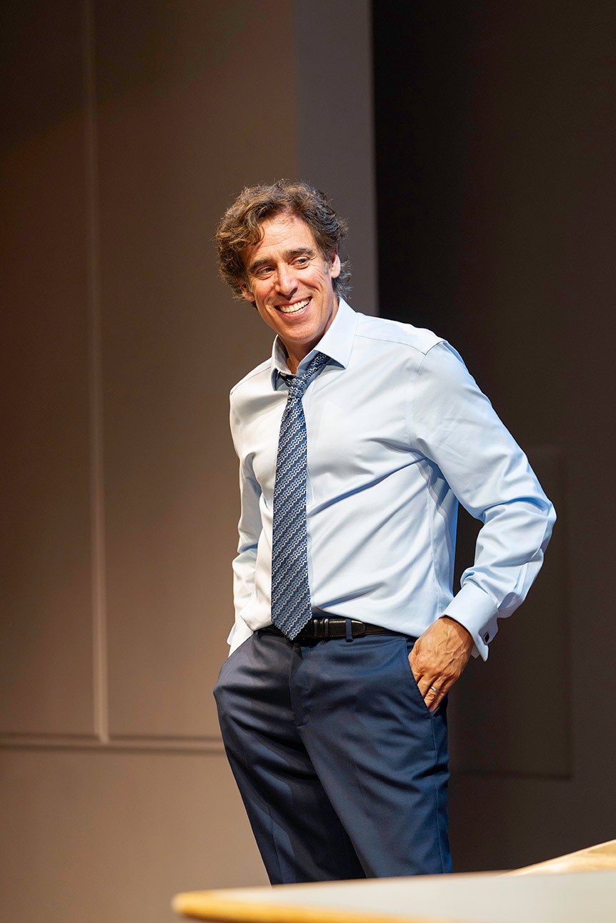 Man in blue shirt and tie smiling, standing with hands in pockets against a plain background.